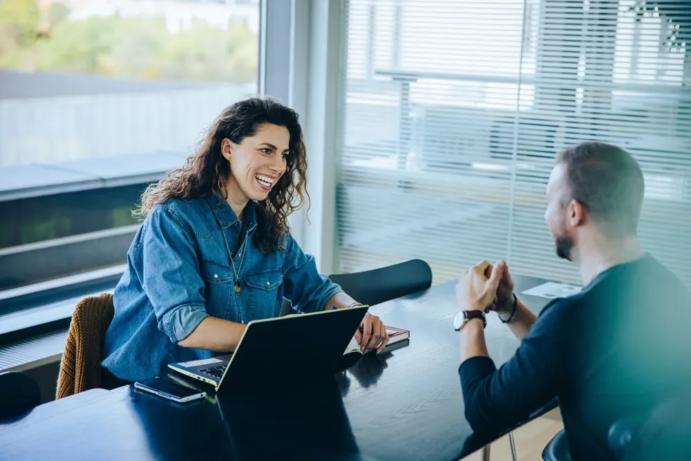 A man and woman sit at a table, focused on a laptop, engaged in discussion or collaboration
